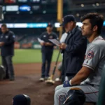 Professional baseball player sitting alone on a dugout bench during a game, looking thoughtful while team staff review paperwork in the background, illustrating designated for assignment in baseball.