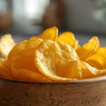 A wooden bowl filled with golden, kettle cooked potato chips showing thick, crispy texture in a softly lit kitchen setting.