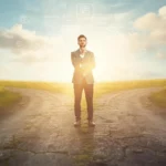 A thoughtful businessman standing at a crossroads where two roads split in different directions, symbolizing “YMMV” (Your Mileage May Vary) and the idea that individual experiences and outcomes can differ.