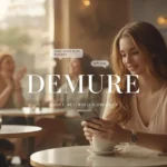 Young woman sitting calmly at a cozy café, softly smiling at her phone while friends laugh in the background, illustrating what does demure mean slang — a modest, reserved, and low-key vibe in modern social media culture.